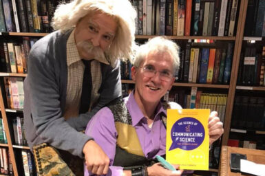 Two men in front of a bookshelf, one is holding a book and the other is dressed as Albert Einstein
