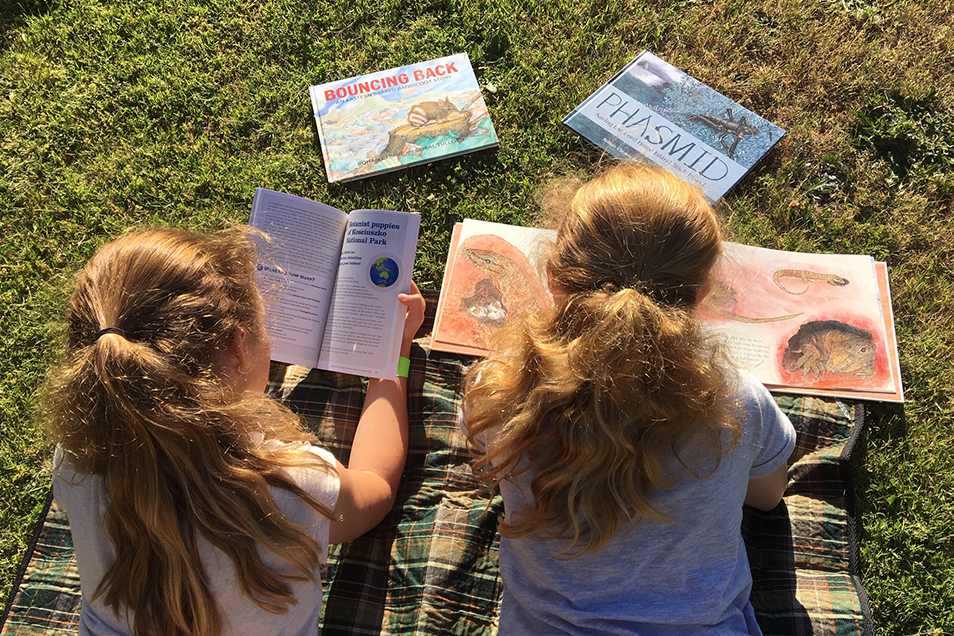Two primary-aged girls laying on their stomachs on grass reading books