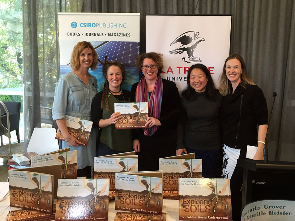 Five women standing together behind a table covered with copies of Exploring Soils, at the launch of Exploring Soils.