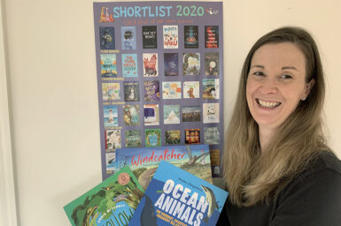 Books Publisher Briana Melideo standing in front of a CBCA 2020 Book of the Year Shortlist poster, holding three CSIRO Publishing children's books
