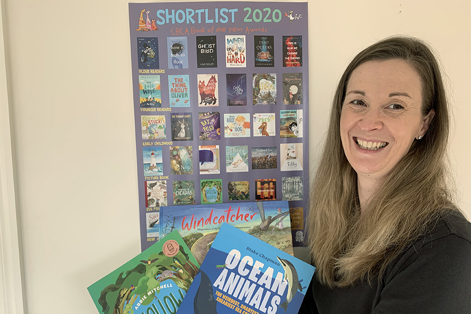 Books Publisher Briana Melideo standing in front of the CBCA Book of the Year 2020 Shortlist poster and holding three CSIRO Publishing books.