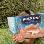 A child laying on grass reading, their face buried in the picture book 'Hold On! Saving the Spotted Handfish'.