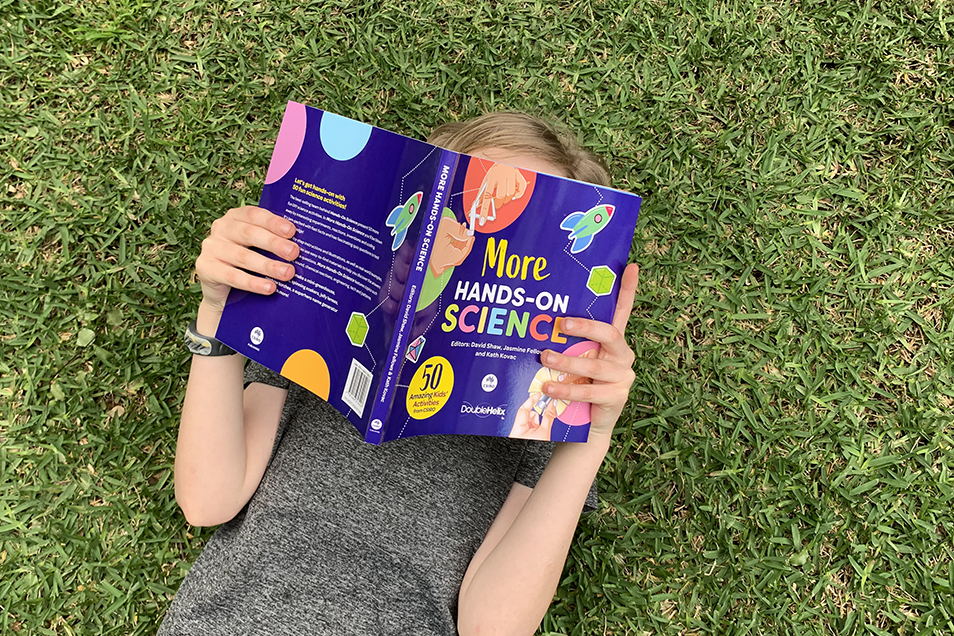A young child lying on their back on grass reading More Hands-On Science. Their face is obscured by the book.