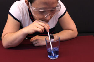 Double Helix editor Jasmine sitting at a table and uses a straw to blow into a glass with blue liquid.
