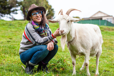 Illustrator Brenna Quinlan crouches in a green field smiling to camera, patting a white goat that stands next to her.