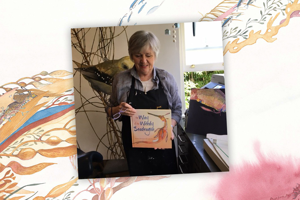 Illustrator Lois Bury standing in her studio surrounded by her art and holding a copy of The Way of the Weedy Seadragon. The photo sits on a background image from the book feautring orange, yellow and pink coloured ocean weeds.