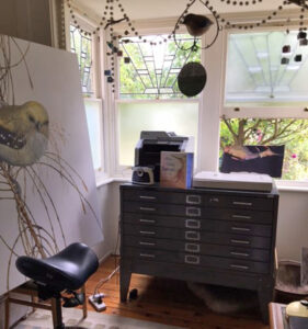 A photo of Lois Bury's studio, including a large painting of a bird, a chest of drawers, and large windows letting in lots of natural light.
