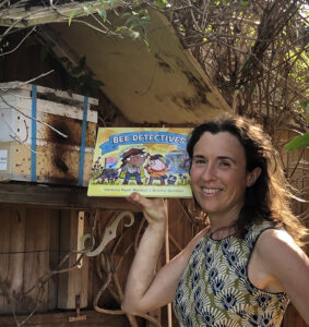 Woman holding a copy of Bee Detectives picture book in front of beehive