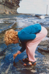 Photo of Ceridwen as a young girl, leaning over a rock pool and poking at something beneath the water. In the distance behind her is a cliff face, and waves breaking against rocks.