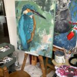 Two paintings of a bird and a koala sitting on easels in an art studio, surrounded by paints, palettes, brushes and a chair.