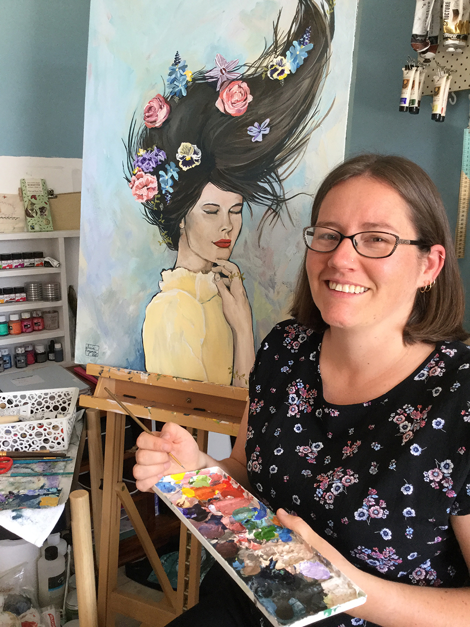 Alicia Rogerson holding a painting palette and sitting in her art studio. She is smiling at the camera and sitting in front of one of her paintings on an easel which depicts a woman with flowers in her hair.
