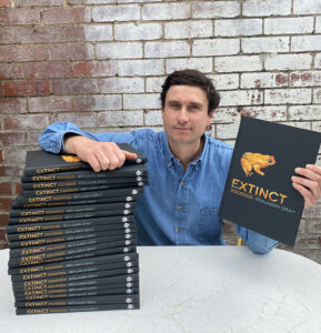 Author Ben Gray seated with a large pile of his book extinct under right arm, and holding a copy to camera in his left.