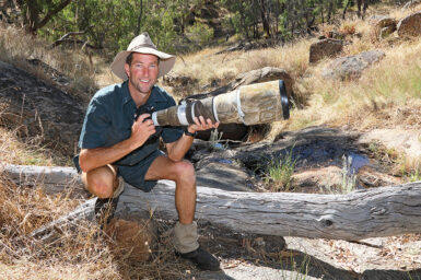 Chris Tzaros sitting on an old fallen log in bushland, smiling and holding a camera with a very long, large lens.