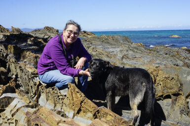 Julie Murphy crouched with her dog on rocks by the ocean.