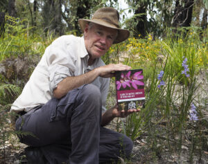 Gary Backhouse kneeling in bushland next to some orchids, smiling and holding his book.
