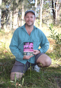 Lachlan Copeland kneeling in bushland next to some orchids, smiling and holding his book.