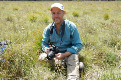 Lachlan smiling and sitting in grasslands, camera poised to photograph some small orchids.