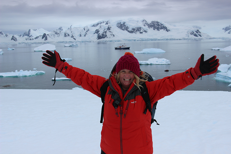 Tracey Gray standing in icy Antarctica, dressed in a red jacket and beanie with her arms raised victoriously in the air.