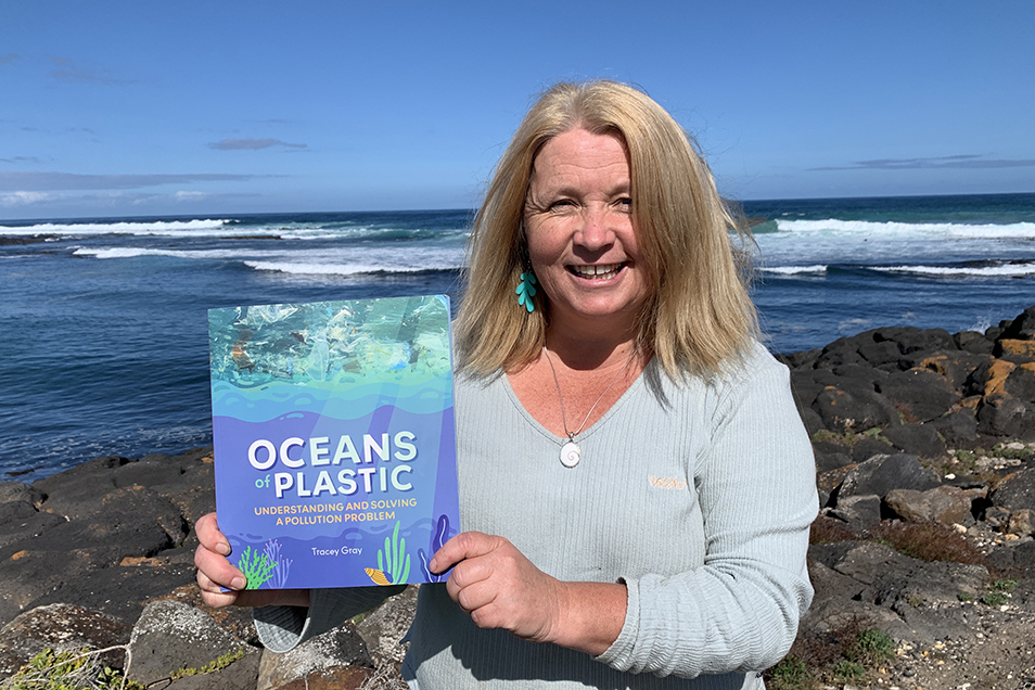 Tracey Gray standing on a rocky seashore, holding a copy of her book Oceans of Plastic.