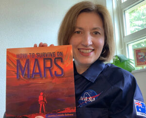 Jasmina wearing blue NASA coveralls, smiling and holding her book 'How to Survive on Mars'.
