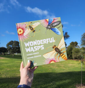 A hand holding the picture book Wonderful Wasps, against a background of green grass and blue sky.