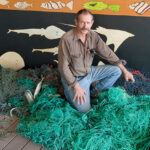 Mike Gillam kneeling in pile of green fishing nets, with artwork of fish and rays behind him.