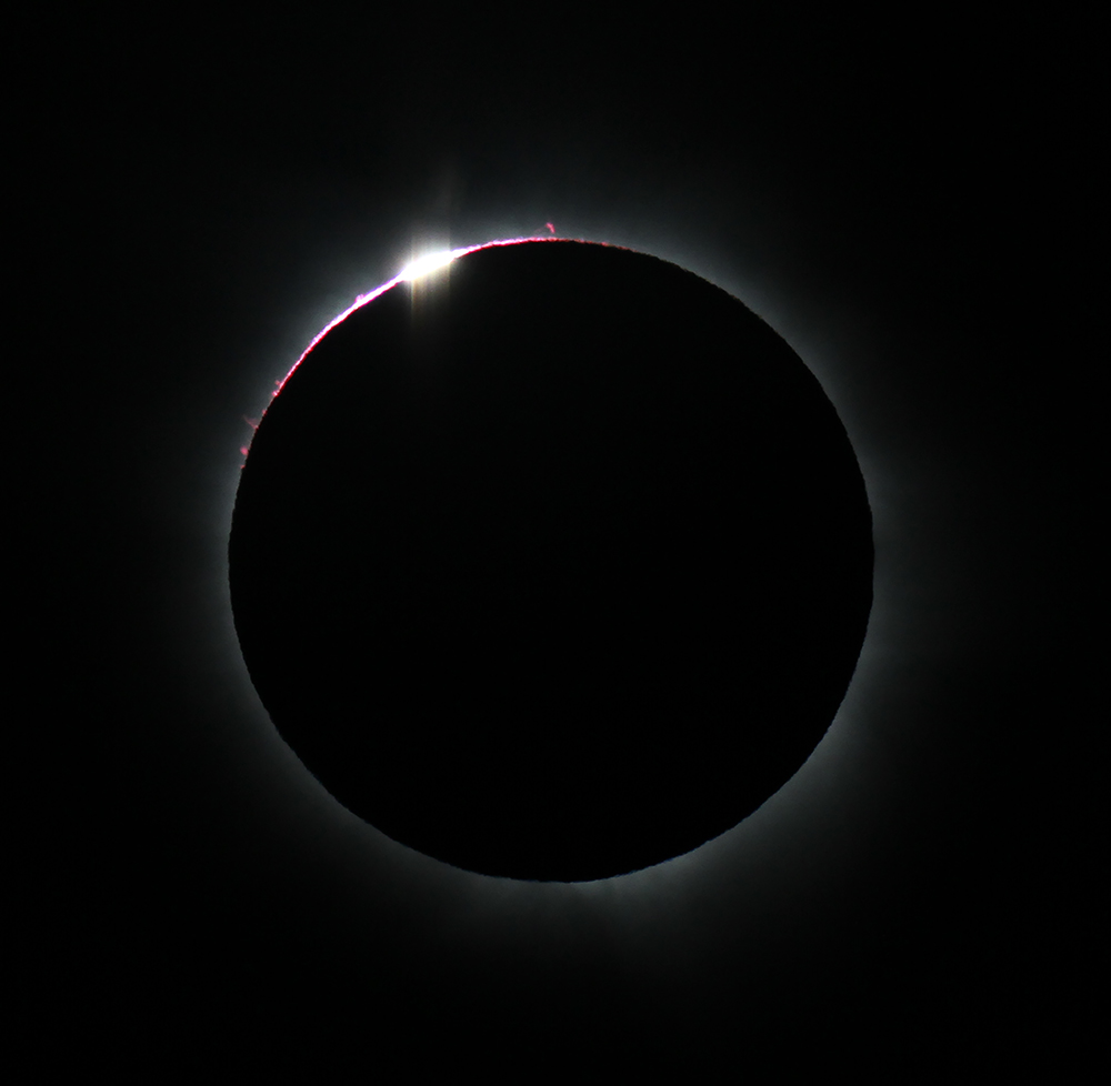 A total eclipse which appears as a black circle with a thin pink and white light around its perimeter against a black sky.