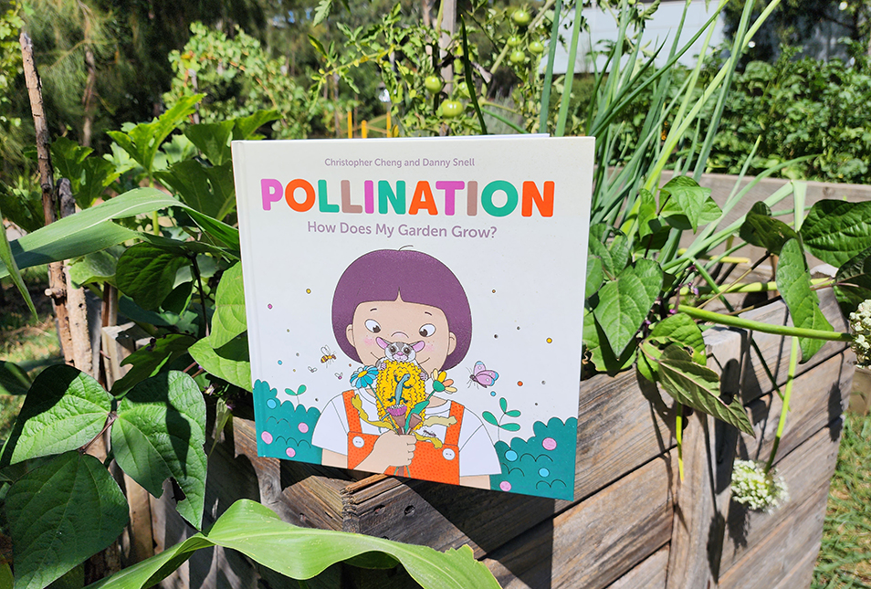 A copy of the book Pollination sitting in a garden box surrounded by leafy green plants.