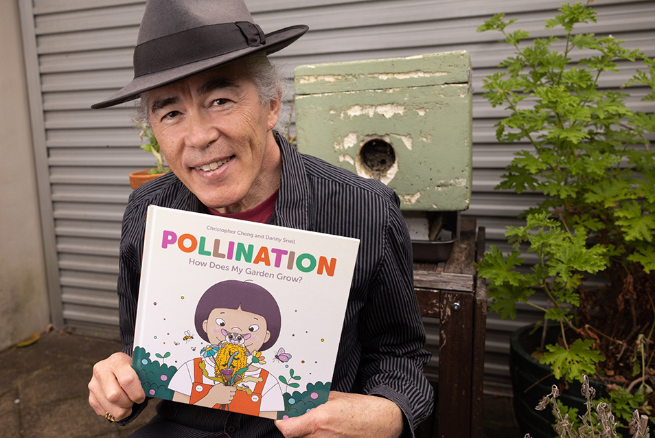 Christopher Cheng smiles at the camera holding a copy of the picture book 'Pollination'. in the background is a native bee hive.