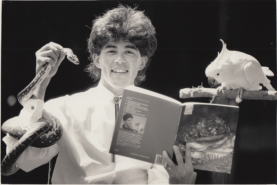 A black and white photo of a young Christopher Cheng holding a snake in his right hand and a copy of a book in his left, while a cockatoo peers over his shoulder.