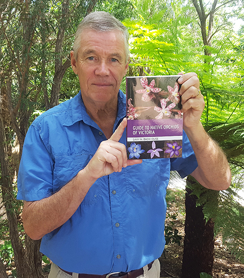 Gary Backhouse holds a copy of A Guide to Native Orchids of Victoria, surrounded by trees.