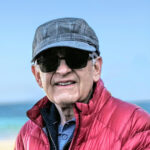 A headshot of author Nick Lomb waring a plaid baseball cap, dark glasses and a red jacket.