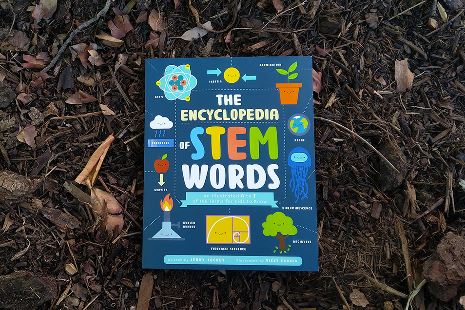 The book 'The Encyclopedia of STEM Words' resting on a bed of dark brown bark chips. The cover is dark blue and features a variety of cartoon-y graphics including a tree, the Earth, an atom, a cloud and a Bunsen burner.