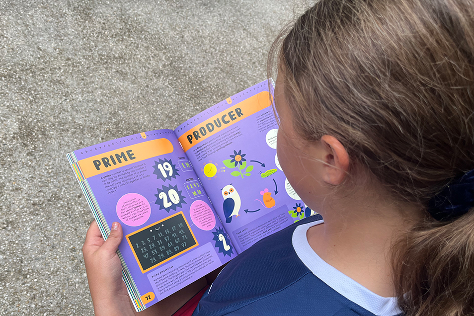 An over-the-shoulder shot of a child with a ponytail reading The Encyclopedia of STEM Words. The book is opened to entries titled 'Prime' and 'Producer'.
