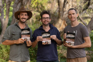 Brendan Schembri, Chris Jolly and Stewart Macdonald standing in a bushland setting, smiling and holding copies of Field Guide to the Reptiles of the Northern Territory.