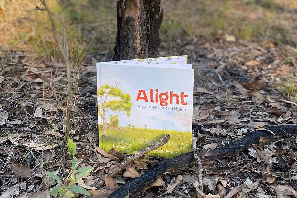 A copy of the picture book Alight, propped up in scrublands with a burnt treetrunk behind it and a charred stick in the foreground.