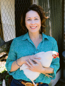 Sam Lloyd smiling and leading against a chook shed. She is holding a white chicken.
