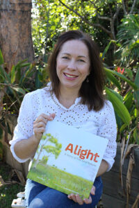 Sam Lloyd in a garden, smiling and holding a copy of the picture book 'Alight'. The cover shows a large eucalyptus tree in grasslands, with smoke rising in the distance.