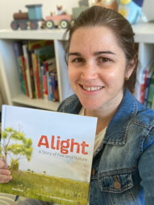 Sam Metcalfe in front of a bookshelf, smiling and holding a copy of 'Alight'.
