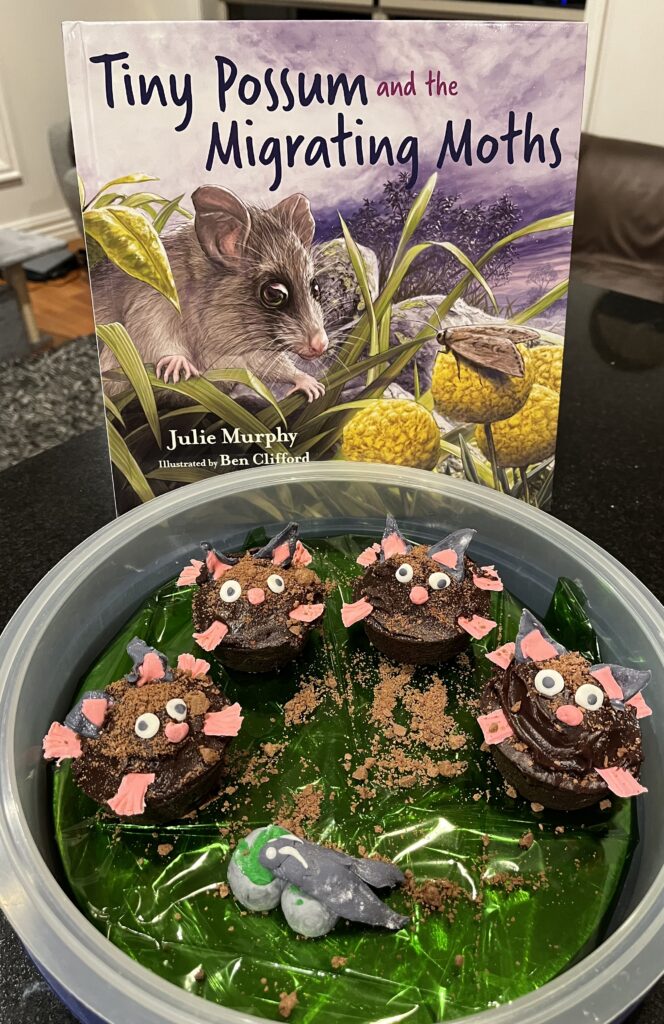 Cupcakes decorated with face, hands and feet to look like a mountain pygmy possum. Behind the cupcakes is the book Tiny Possum and the Migrating Moths, which inspired the cupcakes.
