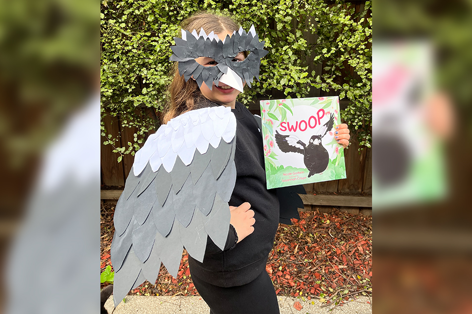A young girl wearing black and white paper magpie wings on her shoulders, a black and white mask on her face and holding a copy of the book Swoop.
