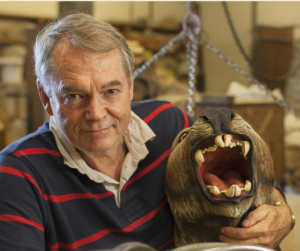 Michael Archer smiles at the camera. His arm os slung around a sculpture of an extinct creature.