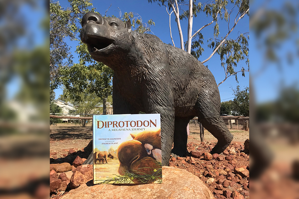 A copy of the children's book Diprotodon in front of a large statue of a diprotodon. They're surrounded by rocky soil and trees.