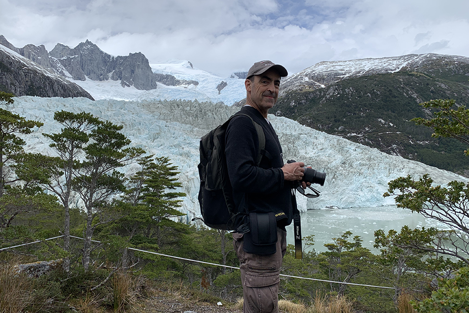 Andrew Plant holds a camera and stands among green plants and trees. Behind him is a large glacier and mountain range.