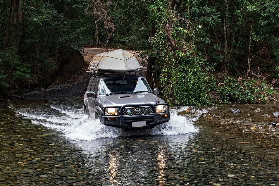 A four wheel drive vehicle with a boat on the roof crossing a creek in a rainforest.