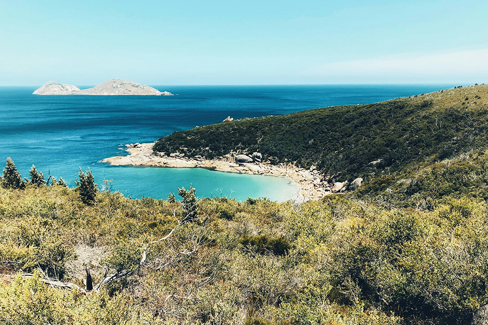 A wide landscape photo of Islands in the distance looking down from the hiking trail at Wilsons Promontory in Victoria, Australia. A rocky shoreline and shrublands are in the foreground.